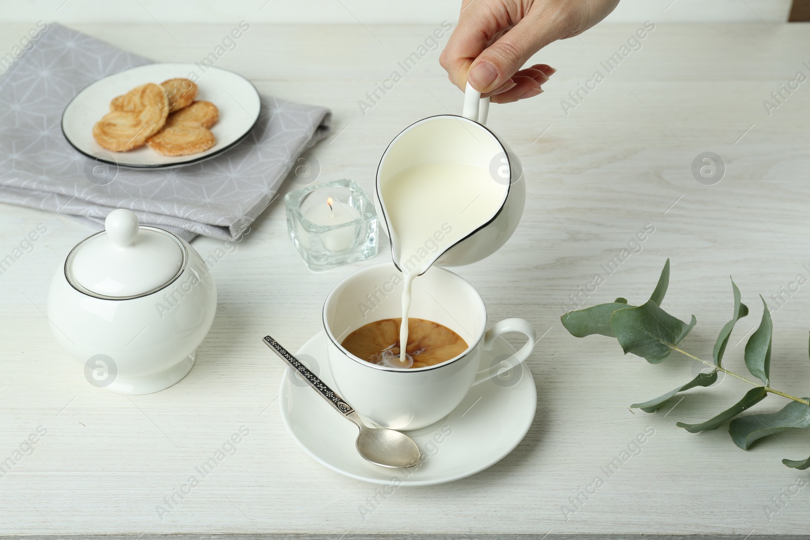 Photo of Woman pouring milk into cup with coffee at white wooden table, closeup