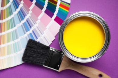 Can with yellow paint, color palette and brush on purple background, flat lay