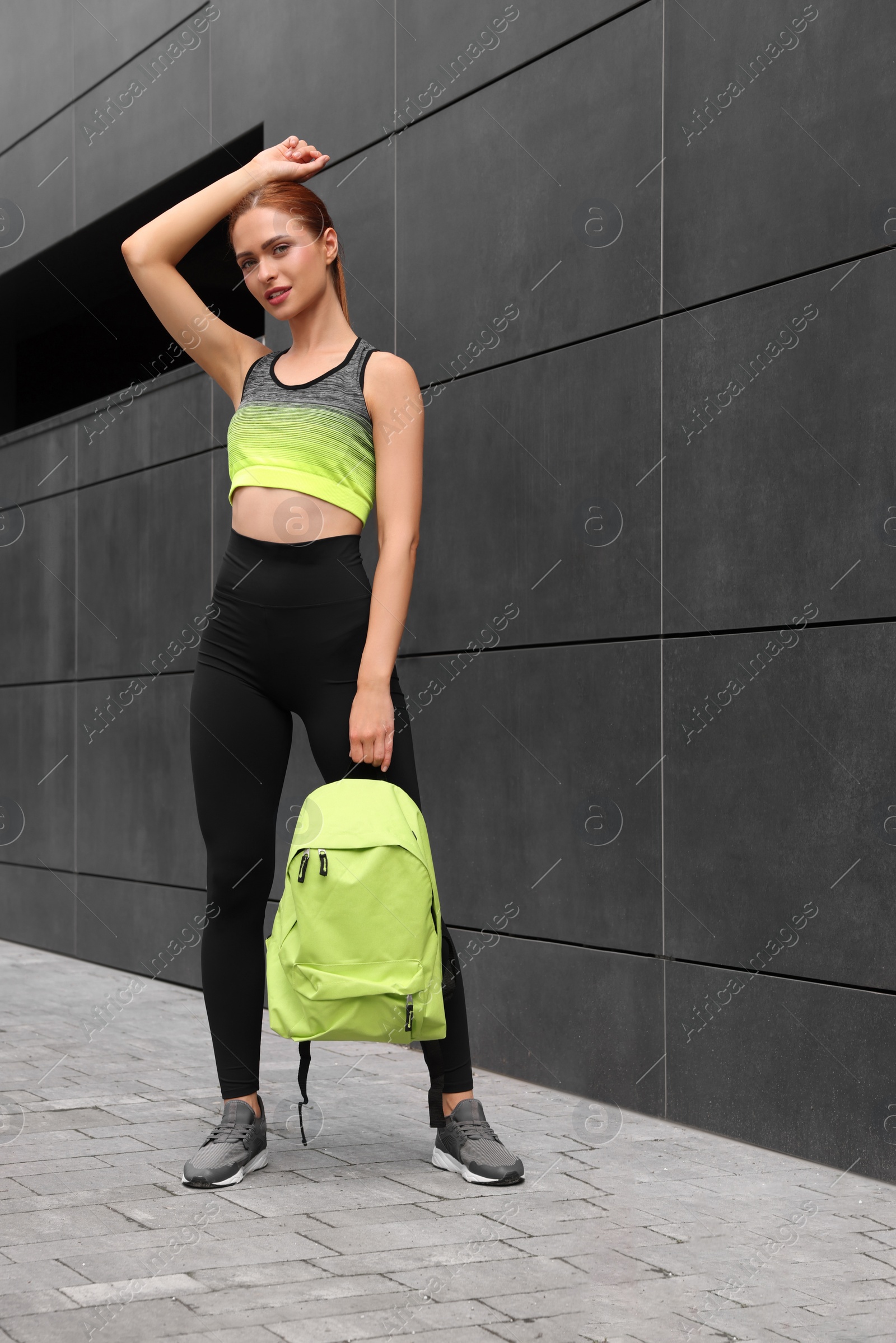 Photo of Beautiful woman in gym clothes posing with stylish backpack near dark grey wall on street
