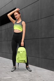 Beautiful woman in gym clothes posing with stylish backpack near dark grey wall on street