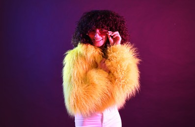 Beautiful young woman in yellow fur coat and sunglasses on color background in neon lights