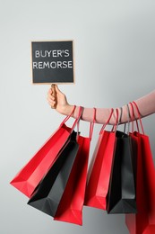 Image of Woman with shopping bags holding small blackboard with text Buyer's Remorse on light grey background, closeup