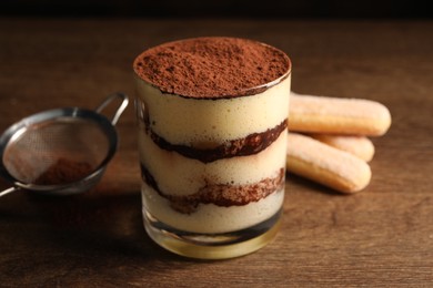 Photo of Delicious tiramisu in glass, biscuits and sieve on wooden table, closeup