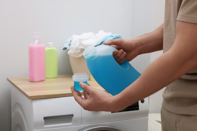 Photo of Man pouring fabric softener from bottle into cap near washing machine indoors, closeup