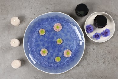 Photo of Plate with water, flowers and burning candles on grey floor, flat lay. Pedicure procedure