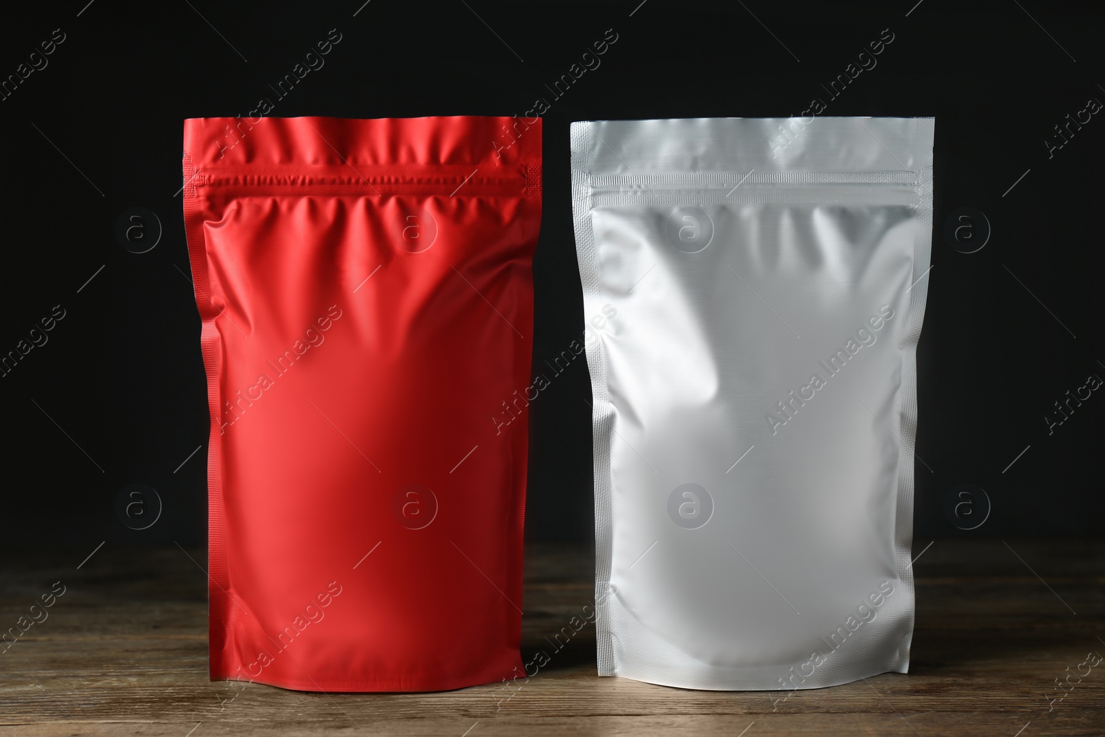 Photo of Different blank foil packages on wooden table against dark background