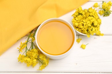 Photo of Rapeseed oil in bowl and beautiful yellow flowers on white wooden table, flat lay