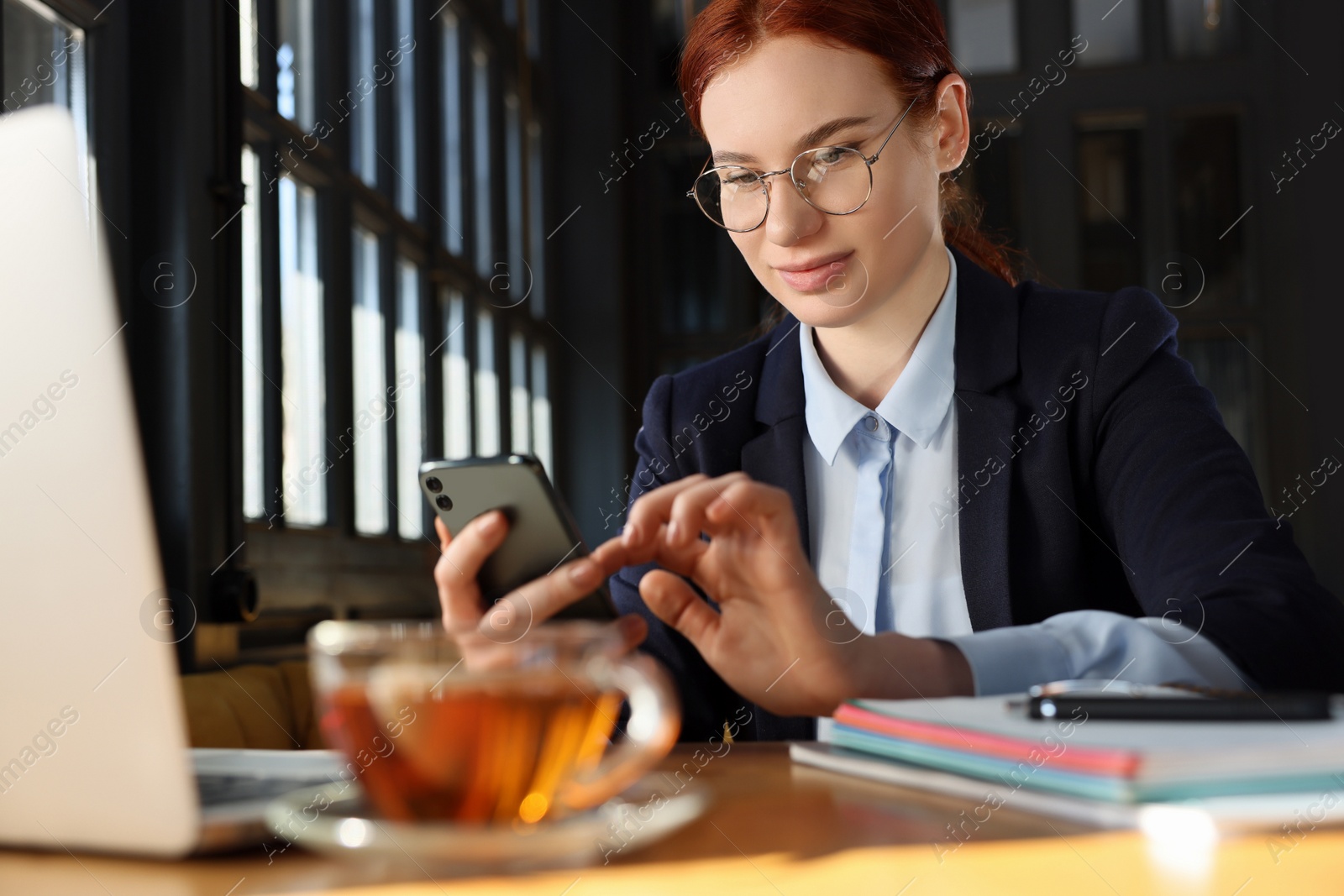 Photo of Young female student with laptop using smartphone while studying at table in cafe