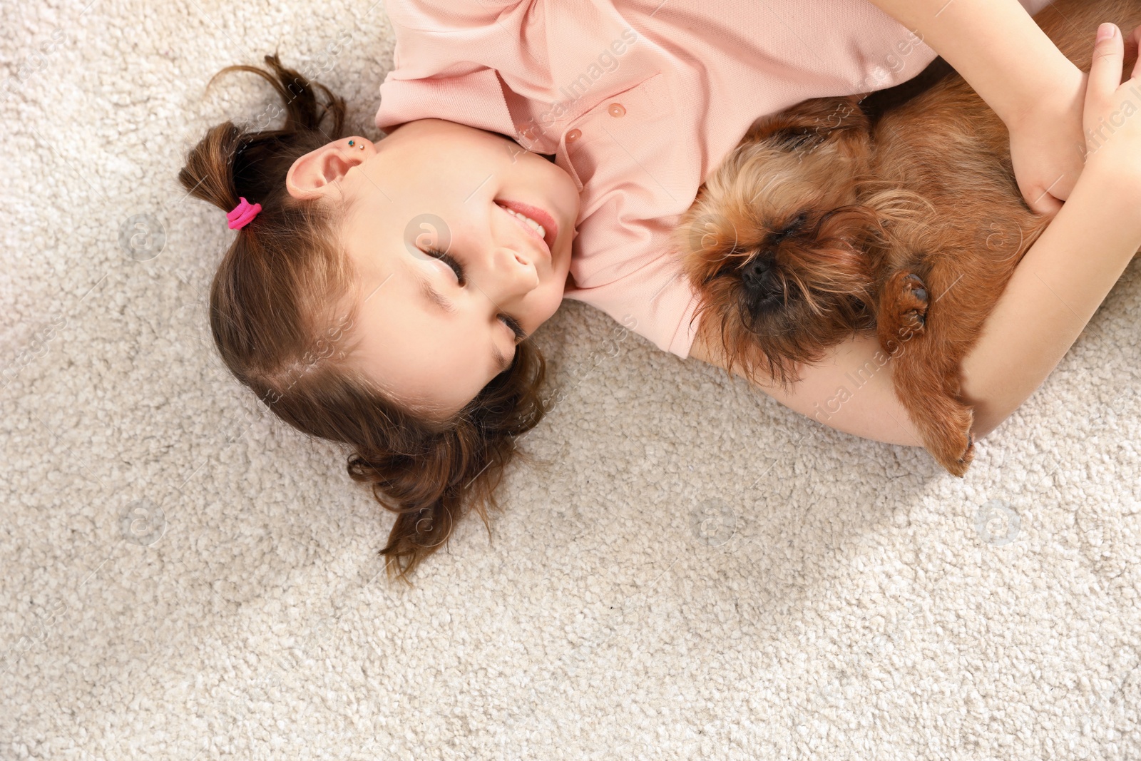 Photo of Portrait of cute girl with funny Brussels Griffon dog lying on carpet, above view. Loyal friends