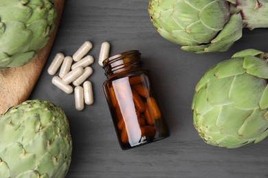 Bottle with pills and fresh artichokes on grey wooden table, flat lay