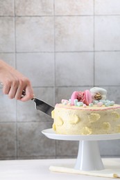 Woman cutting delicious cake decorated with macarons and marshmallows at white wooden table, closeup. Space for text