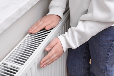 Photo of Girl warming hands on heating radiator indoors, closeup