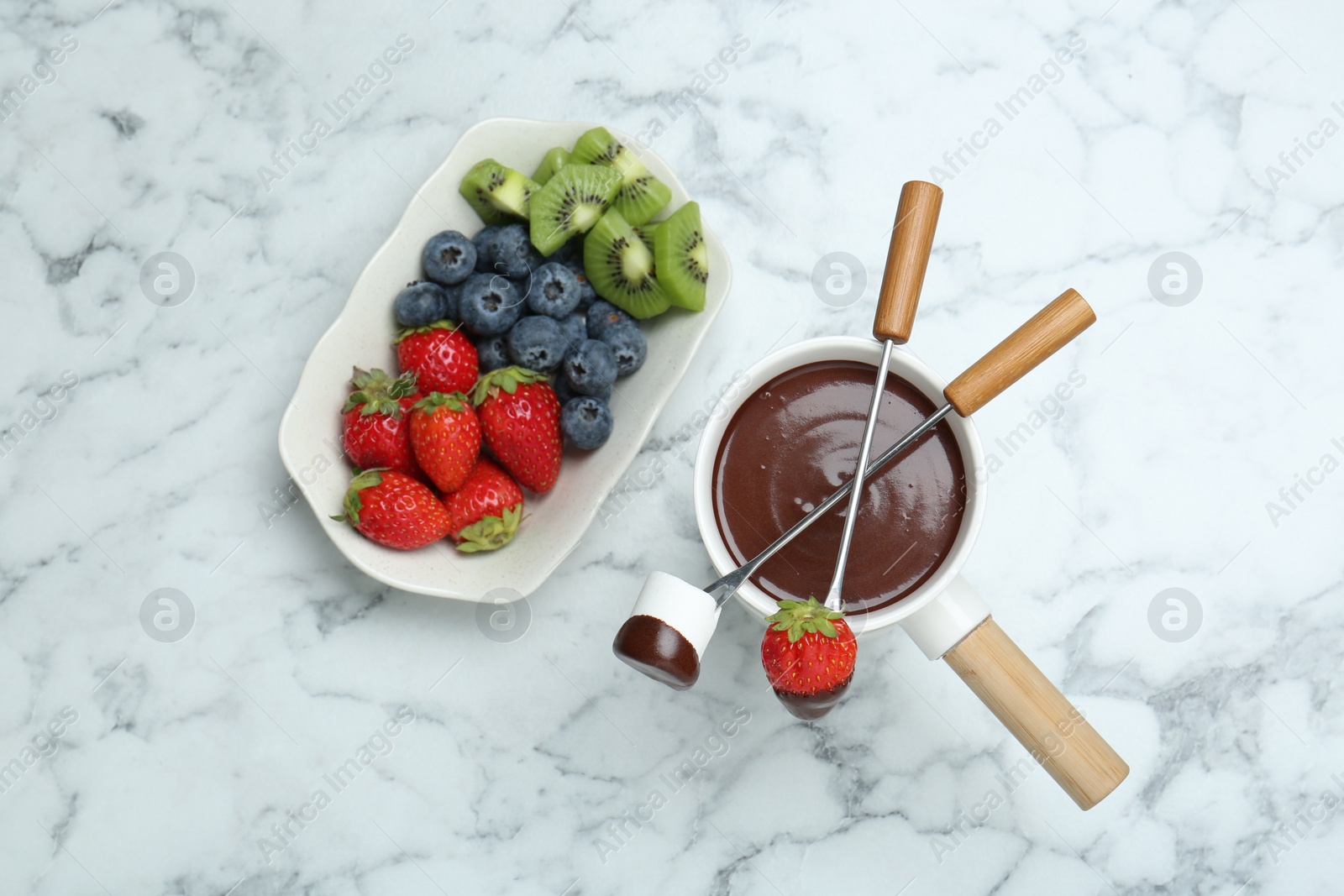 Photo of Fondue pot with melted chocolate, sweet marshmallow, fresh kiwi, different berries and forks on white marble table, flat lay