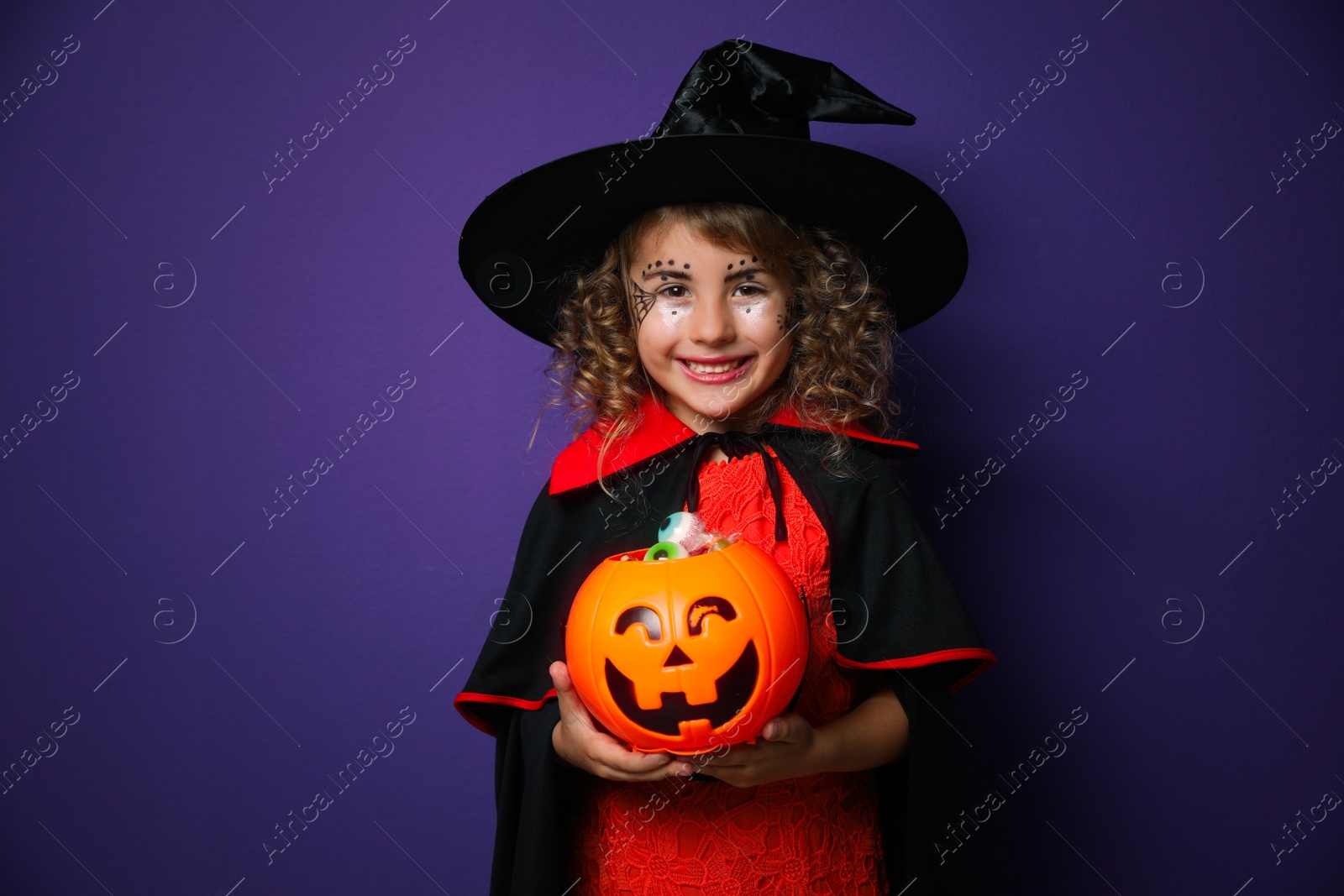 Photo of Cute little girl with pumpkin candy bucket wearing Halloween costume on purple background
