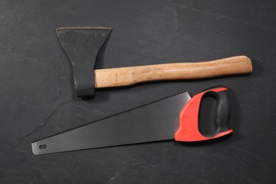 Photo of Saw with colorful handle and axe on black textured background, flat lay