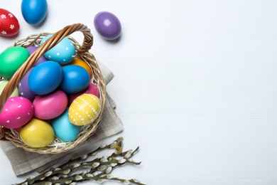 Colorful Easter eggs in wicker basket and willow branches on white wooden table, flat lay. Space for text