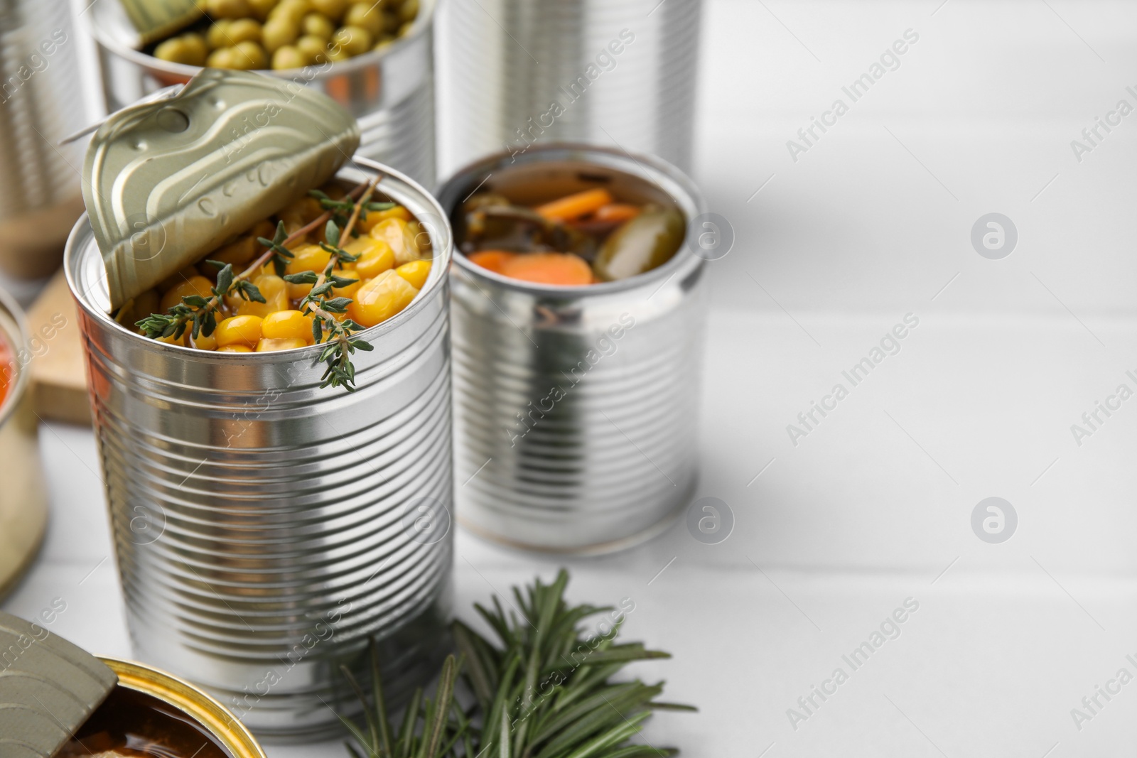 Photo of Open tin cans with corn kernels and different products on white table, closeup. Space for text