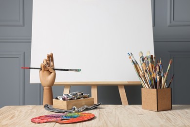 Photo of Easel with blank canvas, hand model and different art supplies on wooden table near grey wall