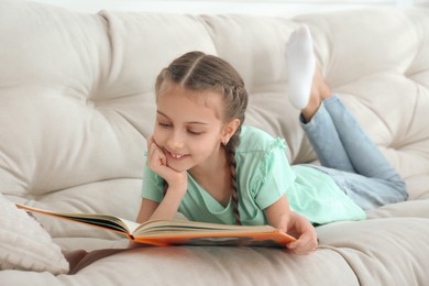 Cute little girl reading book on sofa at home