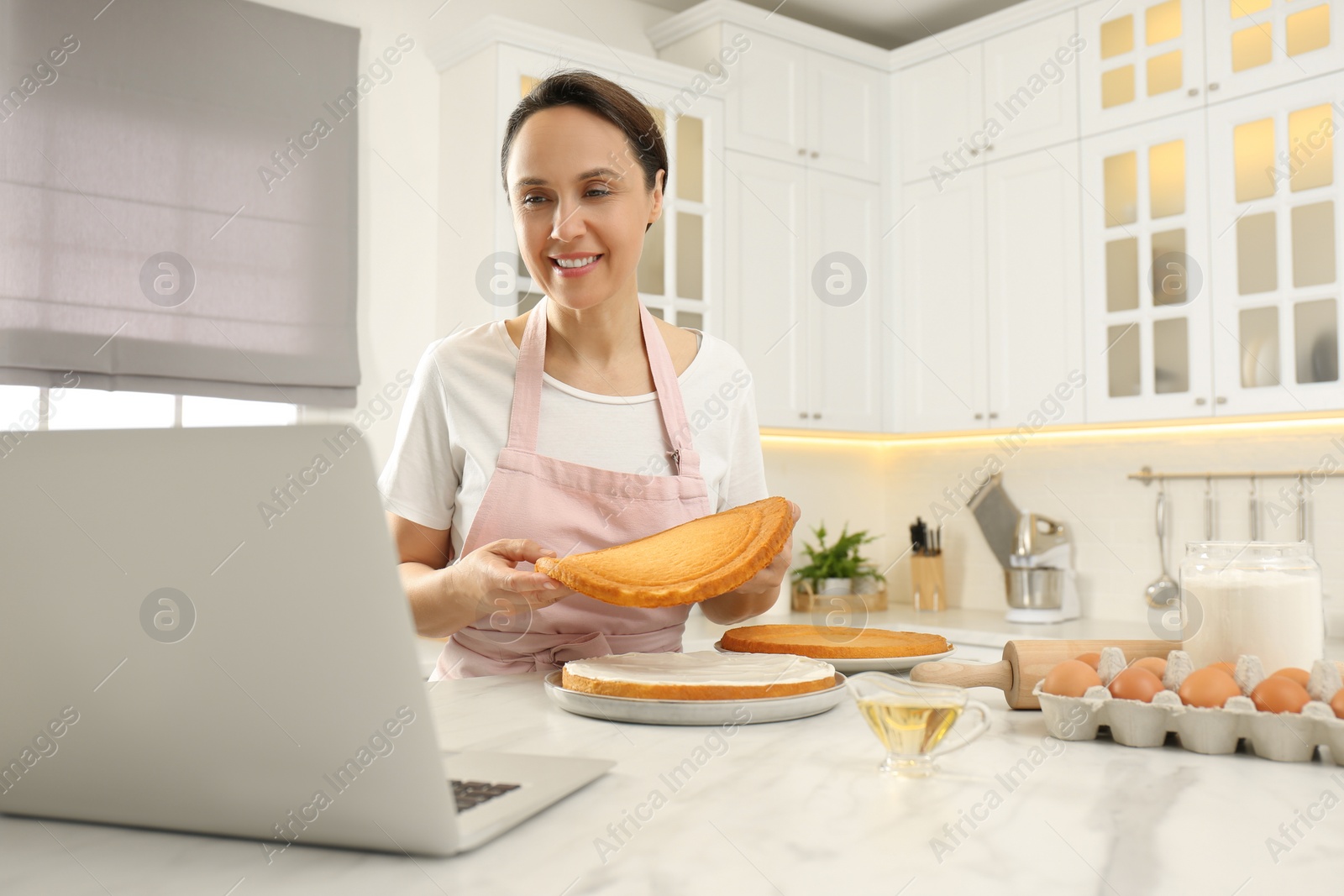 Photo of Woman making cake while watching online cooking course via laptop in kitchen