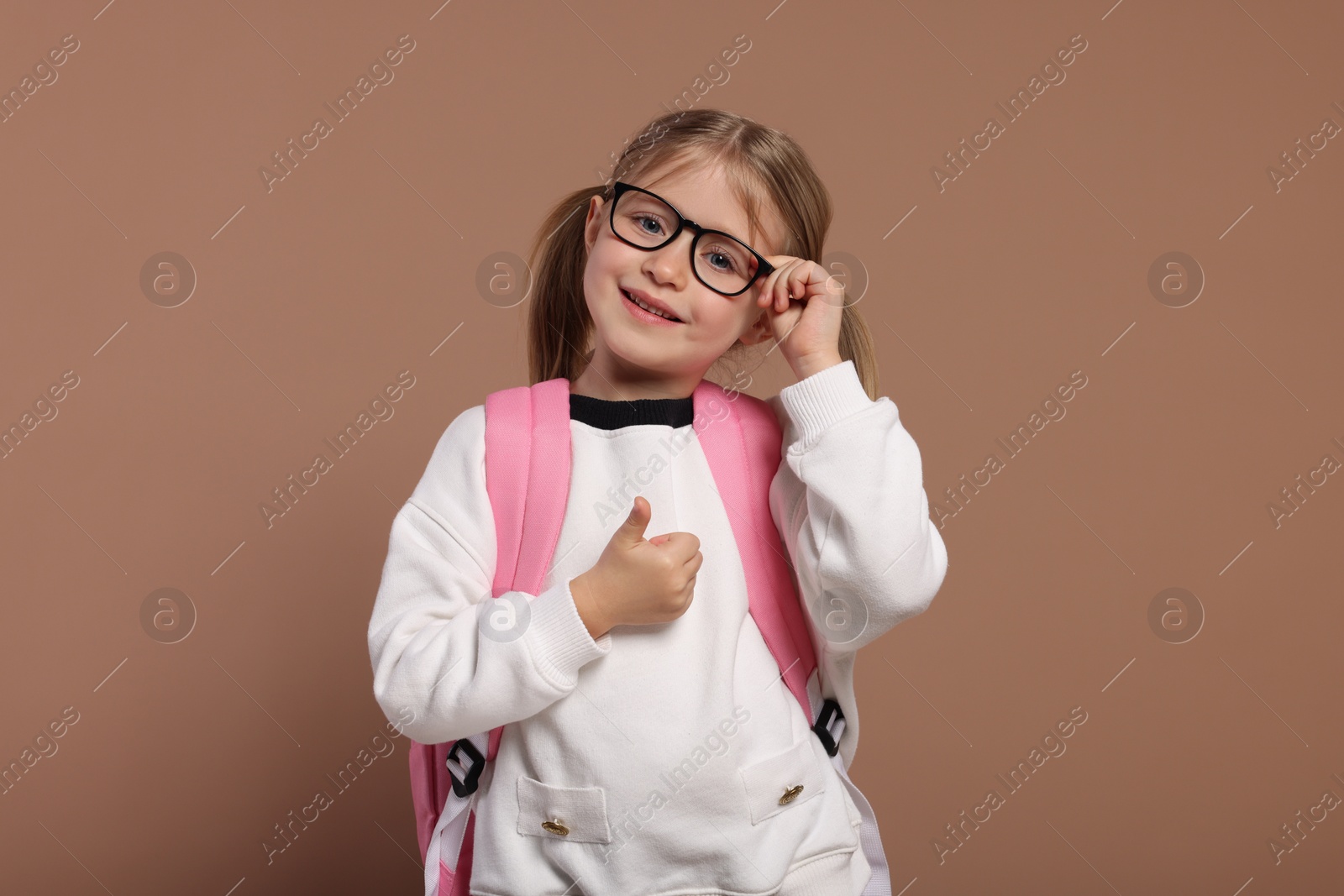 Photo of Happy schoolgirl in glasses with backpack showing thumb up gesture on brown background