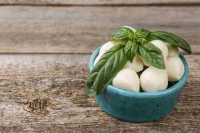 Photo of Tasty mozzarella balls and basil leaves in bowl on wooden table, closeup. Space for text