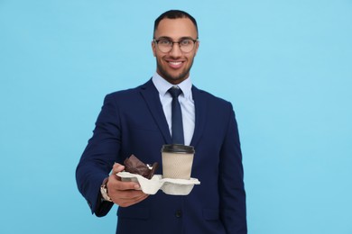 Happy young intern holding takeaway cup with hot drink and muffin against light blue background, focus on cardboard holder