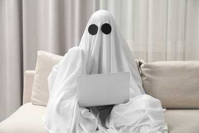 Photo of Creepy ghost. Person covered with white sheet using laptop on sofa at home