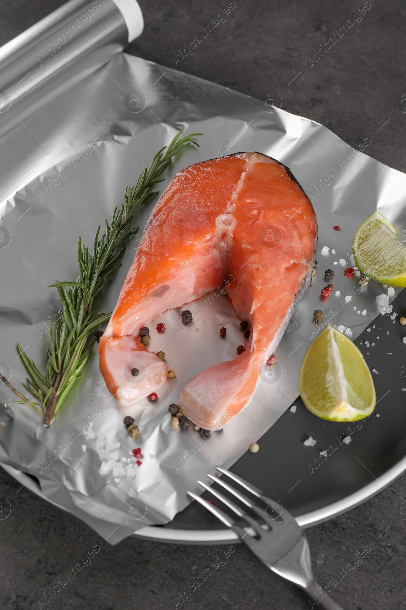 Photo of Aluminum foil with raw fish, lime, rosemary and spices on grey table, closeup. Baking salmon