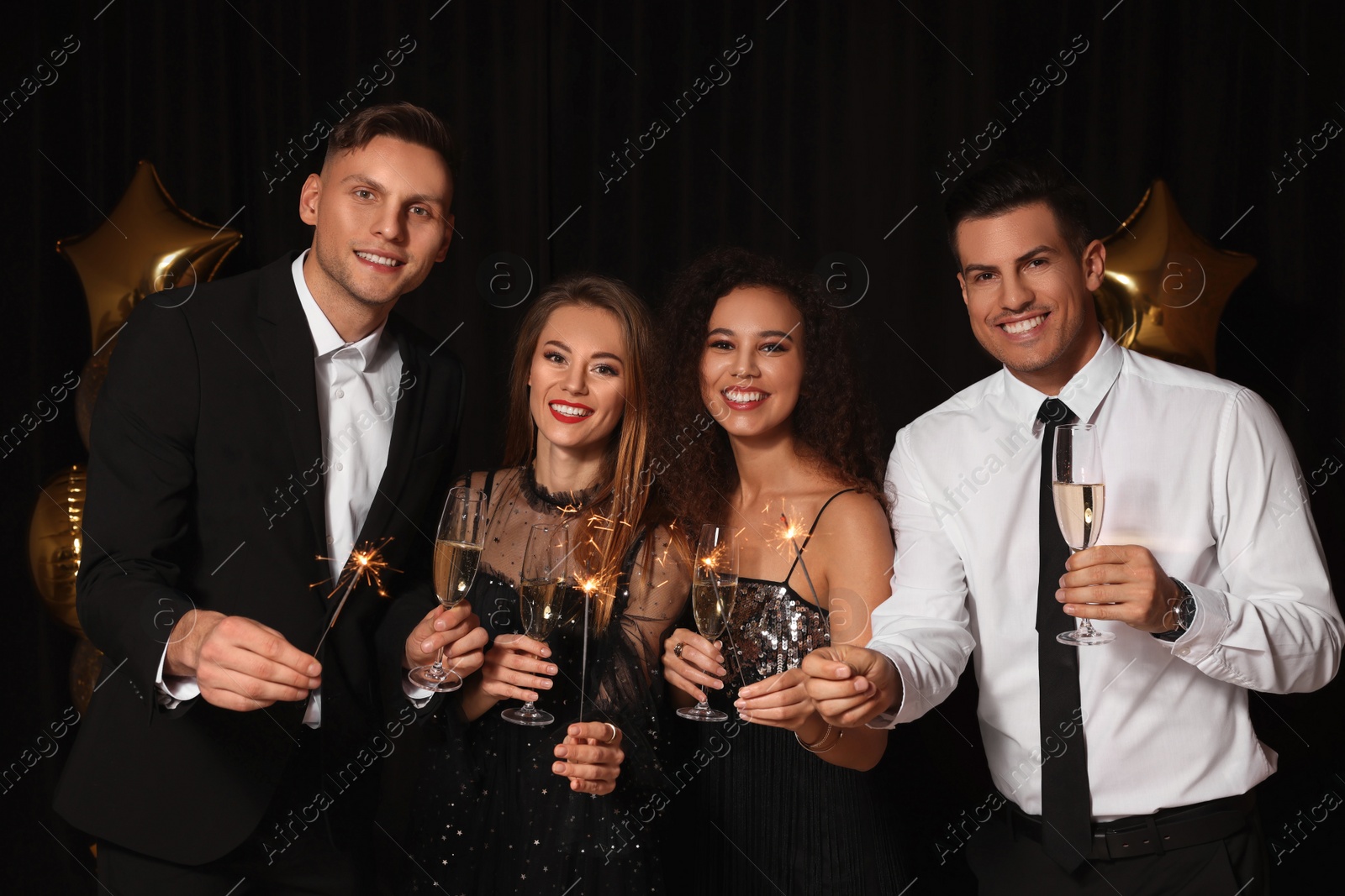 Photo of Happy friends with glasses of sparkling wine and sparklers celebrating New Year on black background