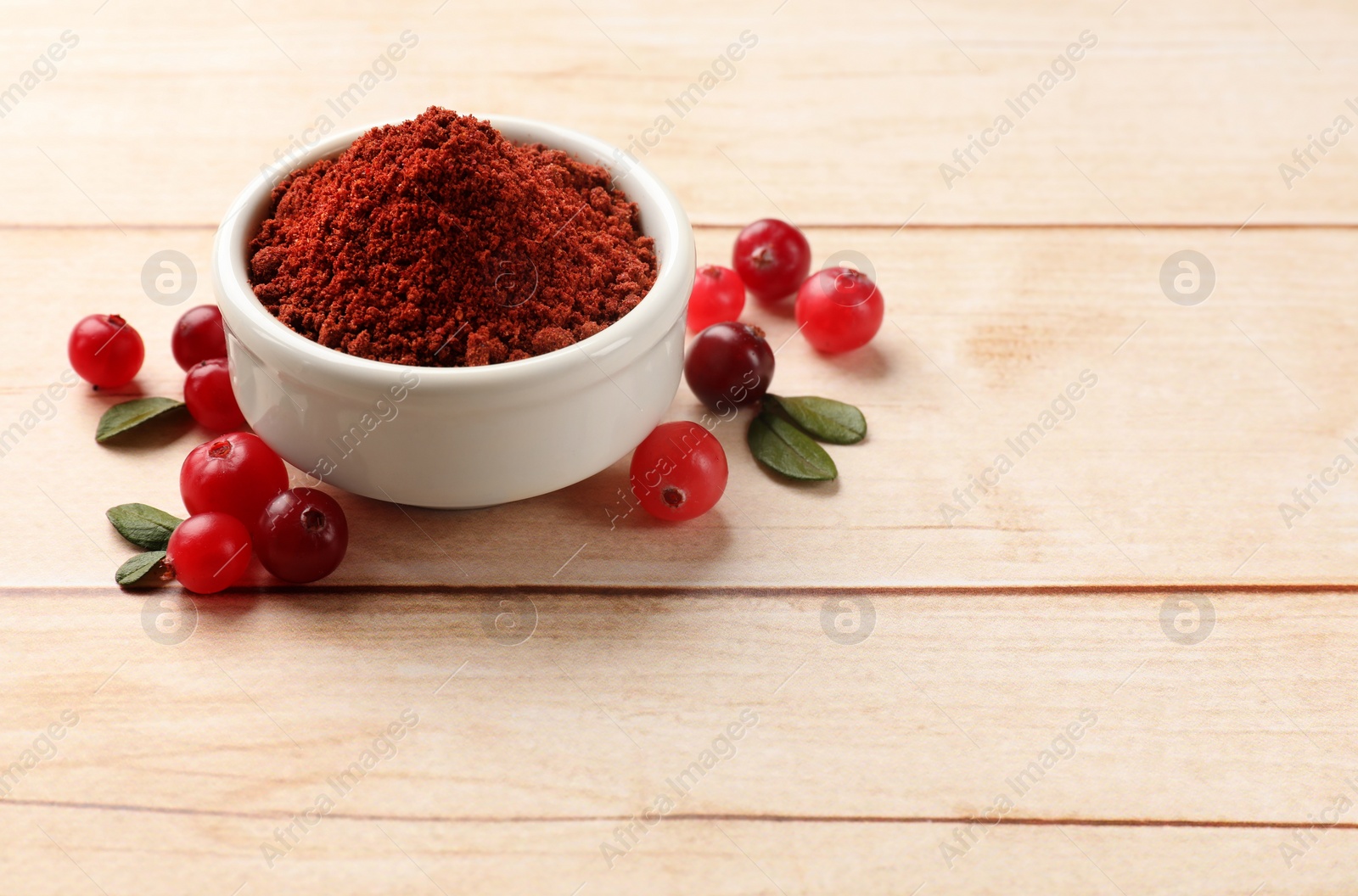 Photo of Dried cranberry powder in bowl, fresh berries and green leaves on light wooden table, space for text