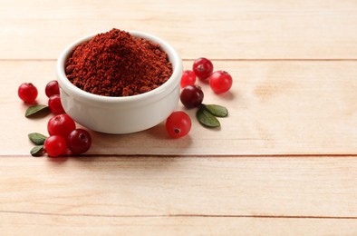 Photo of Dried cranberry powder in bowl, fresh berries and green leaves on light wooden table, space for text