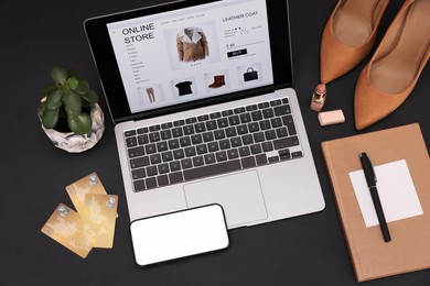 Photo of Online store website on laptop screen. Computer, smartphone, credit cards, women's shoes, stationery and lipstick on black background, above view