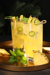 Photo of Glasses of tasty pineapple cocktail with sliced fruit, mint and chili pepper on black table