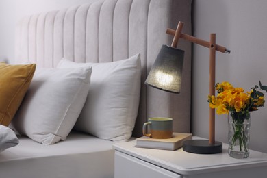 Photo of Stylish lamp, book, hot drink and flowers on bedside table indoors. Bedroom interior element