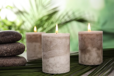 Burning candles and spa stones with palm leaf on table against blurred green background