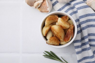 Photo of Fried garlic cloves in bowl and rosemary on white table, flat lay. Space for text