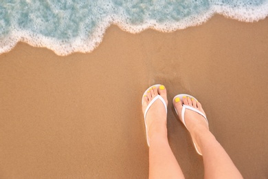 Photo of Top view of woman with white flip flops on sand near sea, space for text. Beach accessories