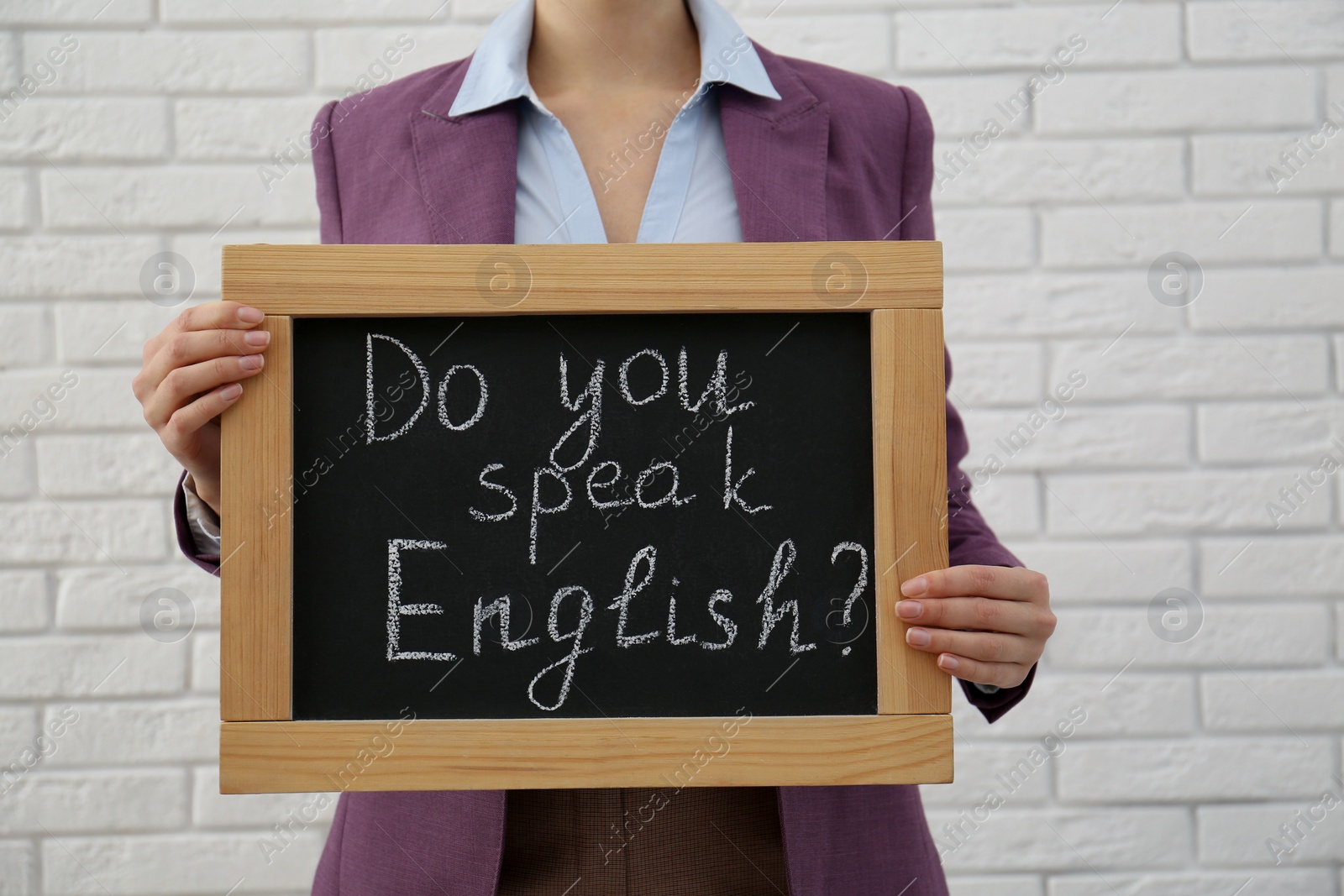 Photo of Teacher holding small chalkboard with inscription Do You Speak English? near white brick wall, closeup