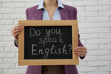 Photo of Teacher holding small chalkboard with inscription Do You Speak English? near white brick wall, closeup