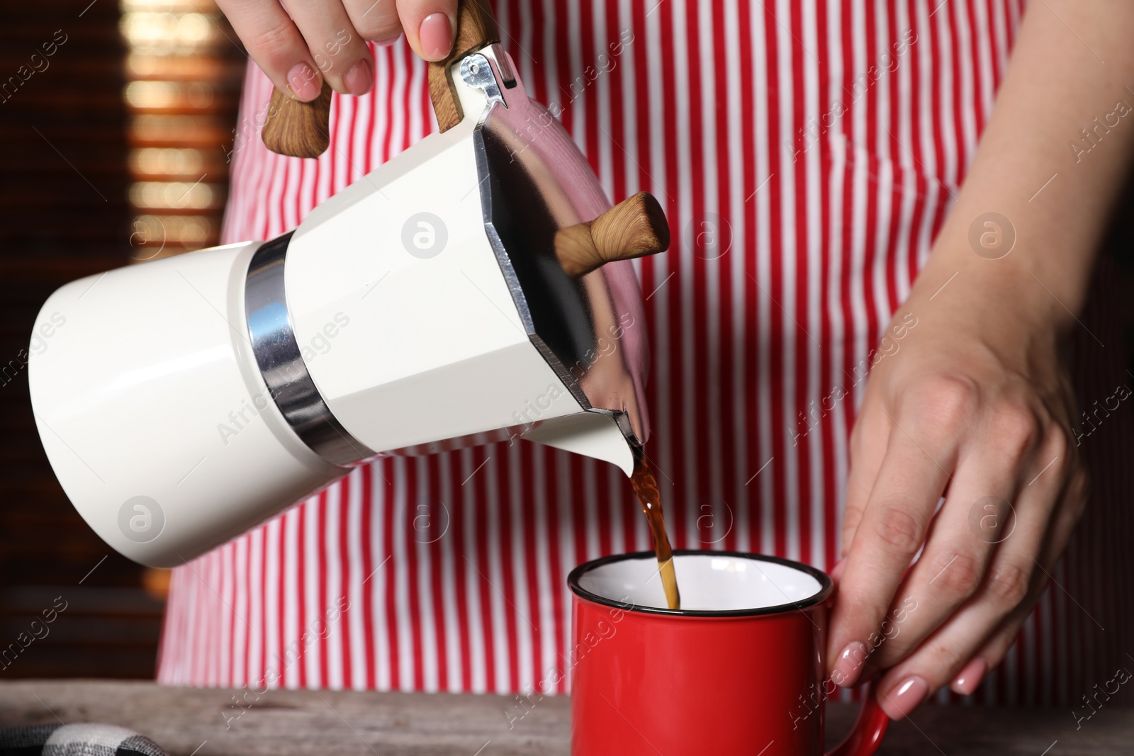 Photo of Woman pouring aromatic coffee from moka pot into cup at wooden table indoors, closeup