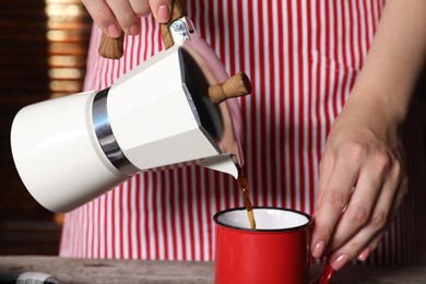 Woman pouring aromatic coffee from moka pot into cup at wooden table indoors, closeup