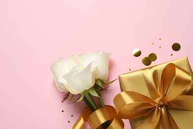 Beautiful golden gift box, flowers and confetti on pink background, flat lay. Space for text