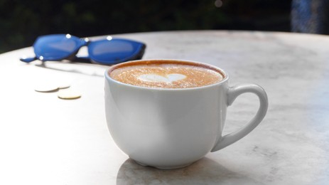 Photo of Cup of delicious coffee and sunglasses on beige marble table outdoors