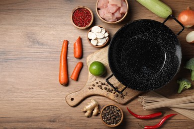 Photo of Empty iron wok surrounded by raw ingredients on wooden table, flat lay. Space for text