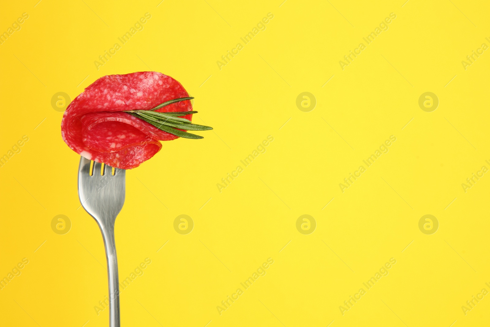 Photo of Fork with tasty slice of salami and rosemary on yellow background, space for text