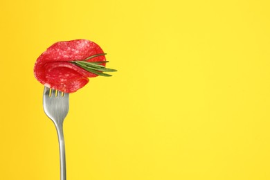 Fork with tasty slice of salami and rosemary on yellow background, space for text