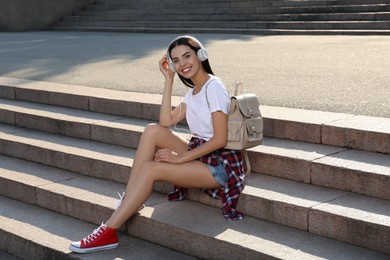 Photo of Beautiful young woman with stylish beige backpack and headphones on stairs outdoors