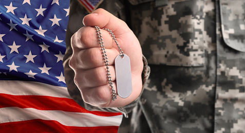 Image of Male soldier holding military ID tag and American flag on background, closeup. Military service