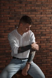 Photo of Young man with tattoos sitting on stool near brick wall